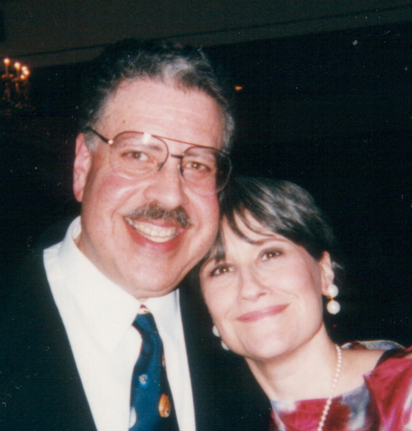 A color photo of a smiling couple, a man and woman. The man on the left has light skin, dark hair and a dark mustache that are graying, and is wearing glasses. The woman on the right has light skin and medium-length dark hair.